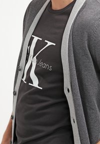 Dunkelgraue Strickjacke mit hellgrauen, gerippten Kanten und Knöpfen, getragen über einem schwarzen Shirt mit weißem Grafikdruck.