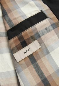 Rutet stof i beige, brun og grå. Designet har kvadrater af varierende størrelse, med en sort sektion og et broderet "NEXT" mærke.