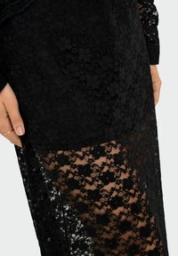 Abito in pizzo floreale nero con pannello trasparente nella gonna, che mostra la pelle sottostante; una mano di colore simile alla pelle con un anello semplice poggia sul lato.
