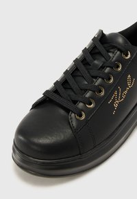 Sneaker en cuir noir avec des œillets dorés, lacets noirs et une texture lisse. Présente un logo stylisé orné de petites clous sur le côté.