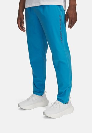 Mężczyzna ubrany w jaskrawo niebieskie sportowe spodnie Under Armour i białe sneakersy, stojący na jednolitym jasnym tle.