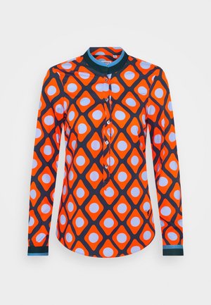 Pitkähihainen napitettava paita, jossa rohkea oranssin ja mustan timanttikuvio, timanttien sisällä valkoiset ympyrät ja tummanvihreä kaulus ja kalvosimet.