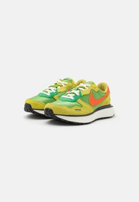 Groene en gele Nike sneakers met witte veters, een oranje logo en een zwarte zool. Suède en stoffen materialen creëren een gelaagd ontwerp.