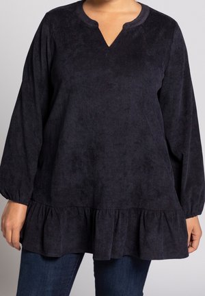 Kobieta nosząca luźną ciemnogranatową bluzkę z długim rękawem, z wycięciem przy dekolcie i falbanką na dole, połączoną z ciemnoniebieskimi dżinsami.