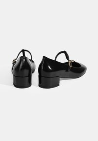 Schwarze lacklederne Schuhe mit abgerundeten Zehen, kleinen Blockabsätzen und dünnen Knöchelriemen, die mit goldfarbenen Hardware-Akzenten verziert sind.