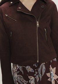 Giacca in suede sintetico bordeaux con design corto, zip argentate e bottoni a pressione. Presenta una chiusura asimmetrica e tasche con zip.