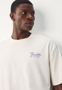 Cream-colored cotton t-shirt featuring a purple Fender logo, short sleeves, and a relaxed fit. Smooth texture and minimalistic design.