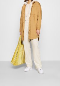 Beżowa kurtka koszulowa, beżowa bluza z kapturem, kremowe spodnie o szerokich nogawkach, białe trampki i teksturowana żółta torba na ramię z subtelnym wzorem.