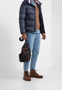 Puffjacka i marinblått med randig krage; brun tröja; ljusblå jeans; bruna snörstövlar; kanvasväska med flera fickor.