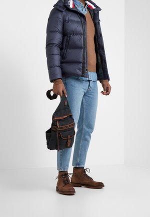 Giacca a vento blu navy con colletto a righe; maglione marrone; jeans azzurri chiari; stivali marroni con lacci; borsa in tela con più tasche.