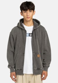 Sudadera gris con cremallera hecha de un tejido suave, que cuenta con un bolsillo tipo canguro, capucha con cordón y una etiqueta de cuero trenzado en la parte frontal.