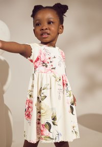 Rochie florală cu bază albă, având imprimeuri cu flori roz și verzi. Mâneci scurte, textură cu nervuri și fustă evazată cu margini cu volane.