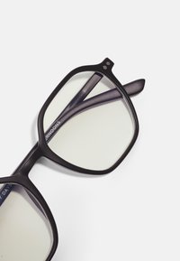 Lunettes noires rectangulaires avec une monture épaisse, finition mate et verres transparents. Accentuées par une branche de temple de couleur violet foncé.