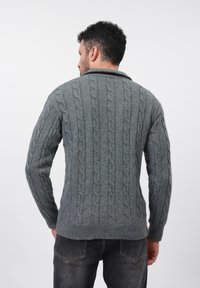 Uomo di spalle che indossa un maglione grigio a trecce e jeans grigio scuro su uno sfondo bianco semplice.