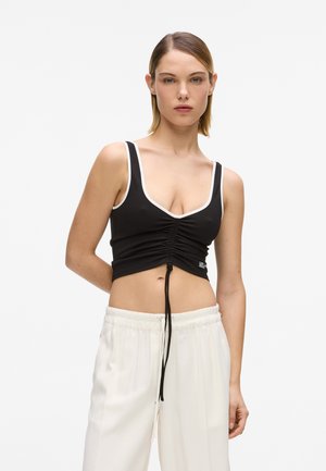 Młoda kobieta w czarnym marszczonym topie z białym obszyciem i luźnych białych spodniach, stojąca na tle jednolitego jasnego tła.