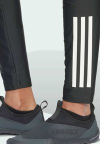 Crne atletske hlače s bijelim trostrukim uzorkom, u kombinaciji s crnim i sivim niskoprofilnim cipelama s teksturiranim gornjim dijelom i s peticama.