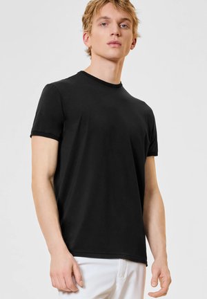 Giovane uomo con una t-shirt nera semplice a maniche corte e pantaloni bianchi, in piedi davanti a uno sfondo grigio chiaro.