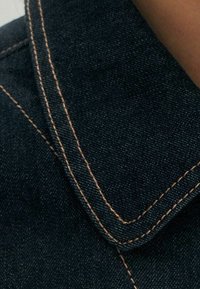 Tmavě modrý denim s výrazně viditelnými dvojitými švy v kontrastní oranžové přízi.