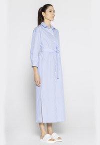 Pasiasta niebiesko-biała sukienka koszulowa z lekkiego materiału, z paskiem do przewiązania, długimi rękawami i rozcięciem do długości kolan. W zestawieniu z białymi sandałami.