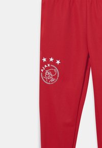 Czerwone sportowe spodnie z gładkiego materiału, z białym logo Ajax oraz trzema białymi gwiazdami ułożonymi w okrągły wzór po lewej stronie.