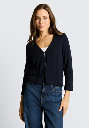 Młoda kobieta o brązowych włosach, ubrana w czarny zapinany na guziki kardigan na białą bluzkę i niebieskie dżinsy, stojąca na tle jasnego, gładkiego tła.