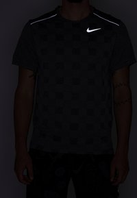 Sportovní tričko s krátkým rukávem Nike v tmavě šedé barvě s kostkovaným vzorem, s reflexními akcenty na ramenou a bílým logem.