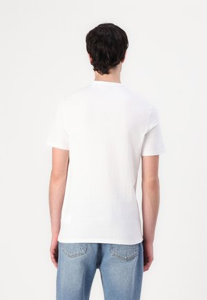 Mann mit dunklen Haaren, der ein schlichtes weißes kurzärmeliges Hemd und blaue Jeans trägt, steht mit dem Rücken zu einem weißen Hintergrund.