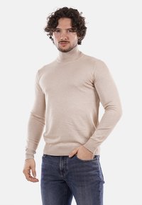 Maglione beige a collo alto lavorato a maglia con maniche lunghe, polsini e orlo a costine, abbinato a jeans blu, modello in piedi contro uno sfondo bianco.