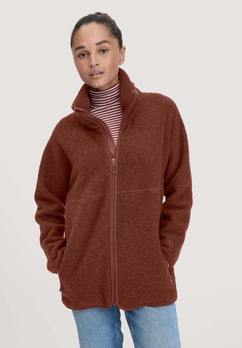 Rustfarbener Fleece-Jacke mit hohem Kragen, Frontreißverschluss und Seitentaschen. Weiche Textur und lockere Passform, getragen über einem gestreiften Rollkragenpullover.