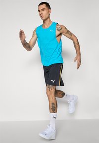 Tirkizna majica bez rukava za sport, tamne kratke hlače s žutim akcentom, bijeli tenisice i bijele sokne do gležnja s logotipom, koje nosi muški model.