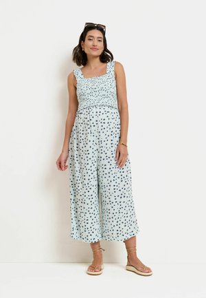 Femme debout portant une combinaison sans manches bleu clair avec un petit motif floral foncé et des sandales beiges avec des brides à la cheville.