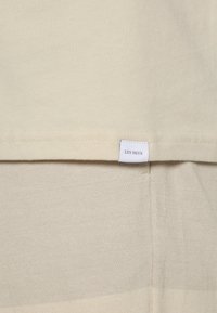 Primer plano de una tela beige con una pequeña etiqueta blanca cosida cerca de la costura que dice "LES DEUX."