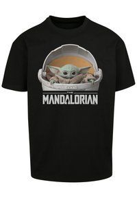 Camiseta negra con un gráfico de un personaje alienígena verde en una cápsula redonda, envuelto en una tela beige. El texto dice "THE MANDALORIAN".