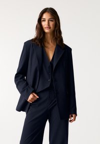 Marineblå jakkesæt med nålestriber bestående af en skræddersyet blazer og matchende bukser, lavet af tekstureret stof. Blazeren har enkelt knappe lukning på brystet.