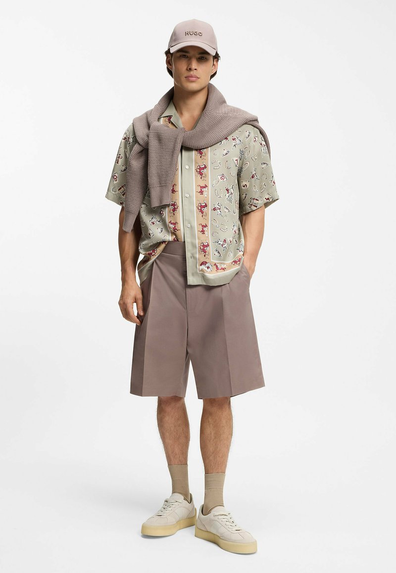 Jeune homme portant une chemise à manches courtes à motifs, un short beige, des baskets, des chaussettes marron clair, une casquette et un pull posé sur les épaules.
