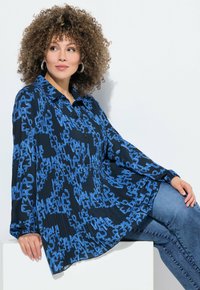 Blusa nera con motivi testuali blu, colletto a camicia, maniche lunghe e design plissettato; abbinata a jeans blu, modello seduto.