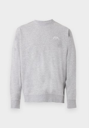 Mees, kes kannab halli dressipluusi, musti lühikeste pükste, mustade sokkide, mustade tossude ja musta pesapalli nokamütsi, seisab ühevärvilise tausta ees.