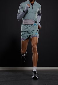 Camiseta deportiva de manga larga en verde menta con cremallera, riñonera gris y pantalones cortos multicolores. Zapatillas de running negras con calcetines blancos, modelo en movimiento.