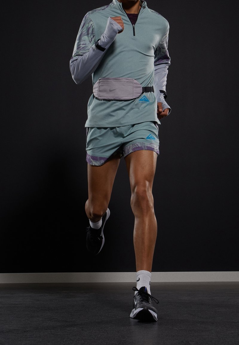 Camiseta deportiva de manga larga en verde menta con cremallera, riñonera gris y pantalones cortos multicolores. Zapatillas de running negras con calcetines blancos, modelo en movimiento.