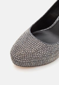 Gros plan d'une chaussure à talon haut argentée scintillante avec des strass cloutés couvrant le bout et la plateforme, doublure intérieure noire.
