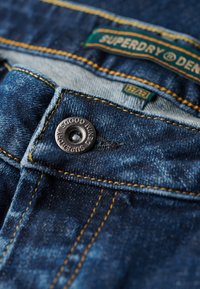 Vaqueros de mezclilla de color azul oscuro, con costuras amarillas prominentes y un botón metálico con la inscripción "Superdry Denim" grabada. Etiqueta visible.