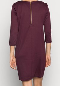 Robe mauve longueur genou avec manches trois-quarts. Comprend une fermeture éclair dorée au dos et une texture de tissu lisse et douce.