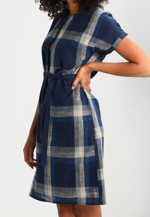 Frau, die ein knielanges, marineblau kariertes Kleid mit kurzen Ärmeln und Taillengürtel trägt, steht mit einer Hand in der Hüfte.