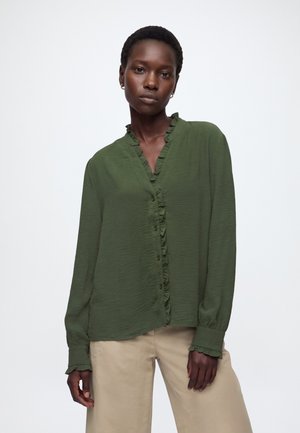 Femme portant un chemisier vert à manches longues avec des détails à volants et un pantalon beige, debout devant un fond blanc uni.