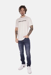 T-shirt bianco a maniche corte con logo nero "ARMANI EXCHANGE", abbinato a jeans blu scuro e sneaker bianche. La vestibilità è casual.
