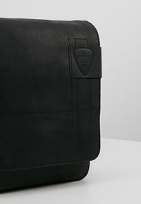 Borsa a tracolla in pelle nera con chiusura a pattina, caratterizzata da dettagli cuciti e una superficie testurizzata. Include un logo del marchio in rilievo.