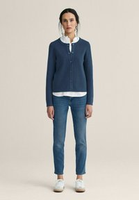 Kvinna stående med en marinblå kofta med knappar över en vit blus, blå jeans och vita sneakers mot en enfärgad beige bakgrund.