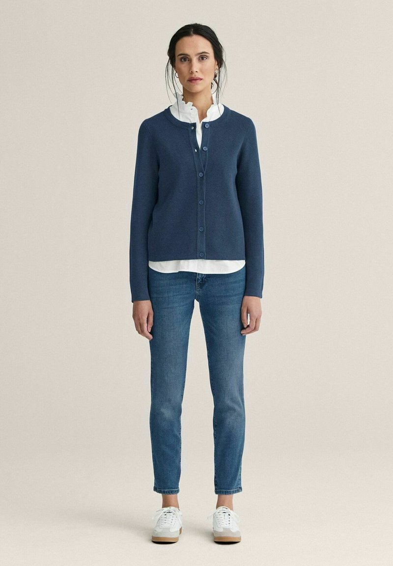 Kvinna stående med en marinblå kofta med knappar över en vit blus, blå jeans och vita sneakers mot en enfärgad beige bakgrund.