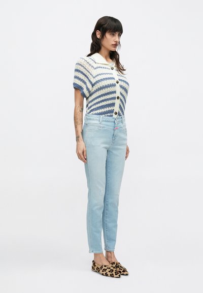 Πλεκτή ζακέτα με κοντά μανίκια σε μπλε και κρεμ ρίγες, με μαύρα κουμπιά· συνδυασμένη με ανοιχτόχρωμα μπλε τζιν και παπούτσια με λεοπάρ μοτίβο.