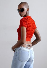 Top corto texturizado rojo con mangas cortas, combinado con jeans azul claro que cuentan con un cinturón decorativo. Diseño suave y ajustado con detalles prominentes.